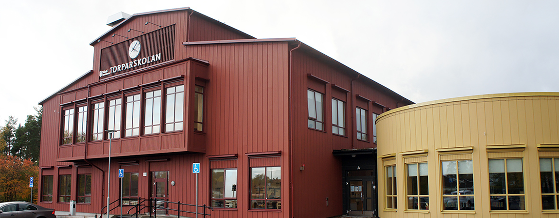 Röd träbyggnad, med gul mindre träbyggnad bredvid. På den röda fasaden står det Torparskolan i vita bokstäver och hänger en stor klocka på fasaden