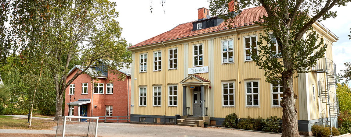 Gul träbyggnad med grön dörr. På fasaden sitter en skylt med texten Öjaby skola