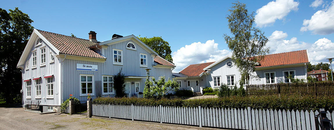 Ljusgrå träbyggnad, på fasaden sitter en skylt med texten Ör skola. Framför skolan en trädgård med staket runt om 